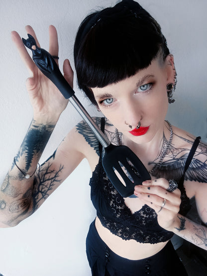 Missjescho, tattooed with short black hair and red lipstick holds the Alchemy black bat shaped silicone spatula with a metal handle, wearing a black lace top against a plain light background.