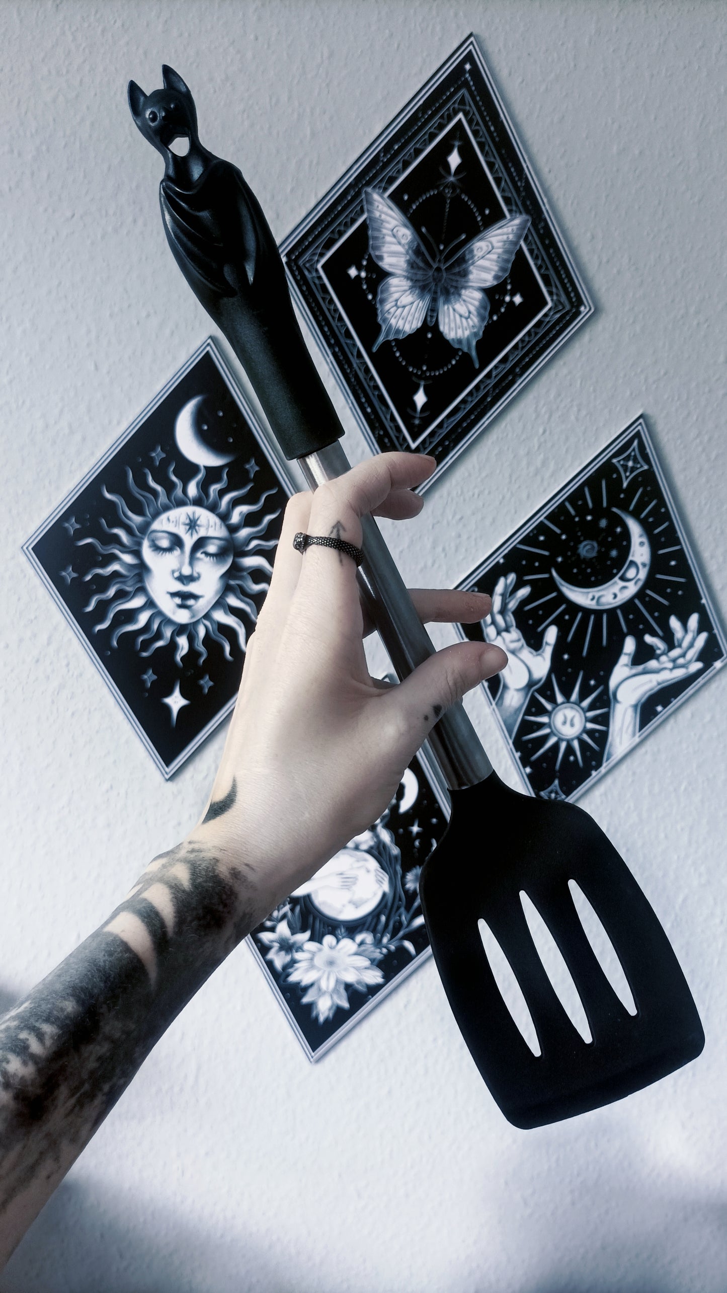 A tattooed hand holds a black spatula with a bat-shaped handle against a white wall, with four black tarot card prints featuring celestial and mystical designs arranged behind it.