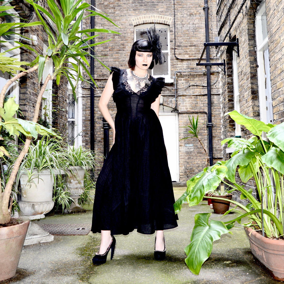 A woman stands in a brick courtyard wearing a long black vintage-style dress with ruffled sleeves, a sheer bodice, and a feathered headpiece. Large potted plants frame the scene.