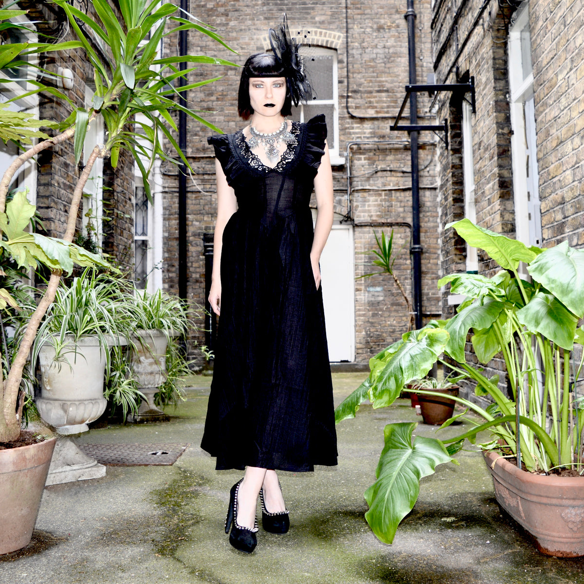A woman modeling a black 1920s-style flapper dress with ruffled sleeves, a sheer mesh bodice, and a matching feather fascinator, standing in an outdoor courtyard with potted plants and brick walls.