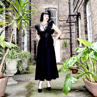 Julia Rose stands in an outdoor courtyard between brick buildings and potted plants, wearing a long black goth dress with ruffled sleeves, a sheer embroidered bodice, and black heels.