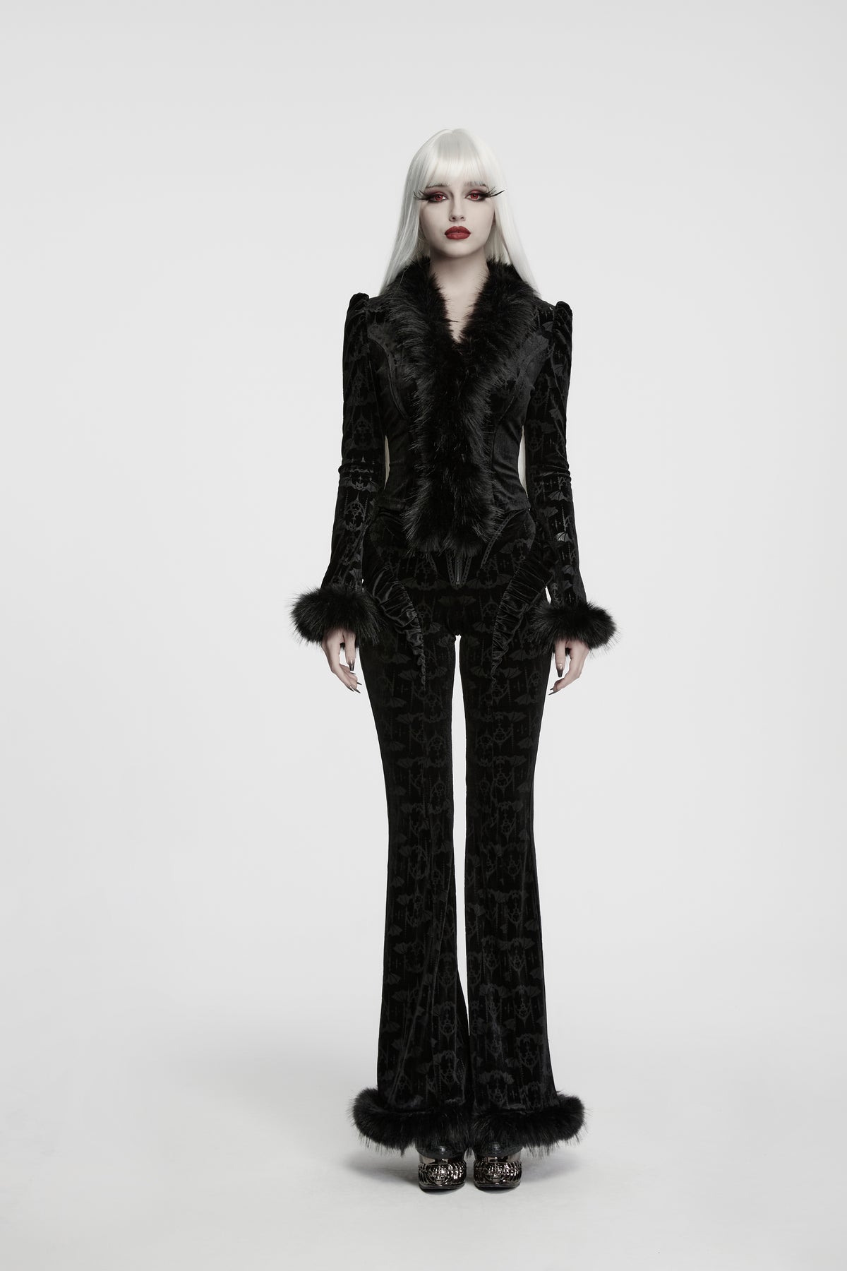 An alternative model with platinum blonde hair wearing a black bat patterned velvet jacket and flared trousers with a black fur collar and cuffs, photographed against a seamless white studio background.