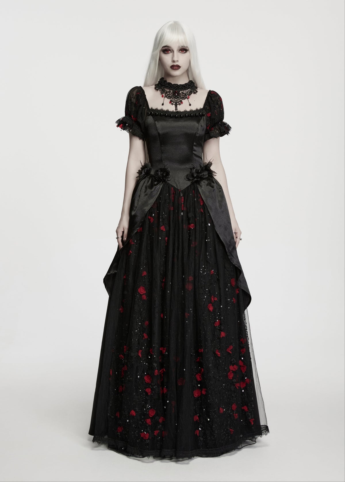 A gothic-style black wedding gown with red rose embroidery, modelled by a person with platinum blonde hair and pale makeup against a light grey studio background.