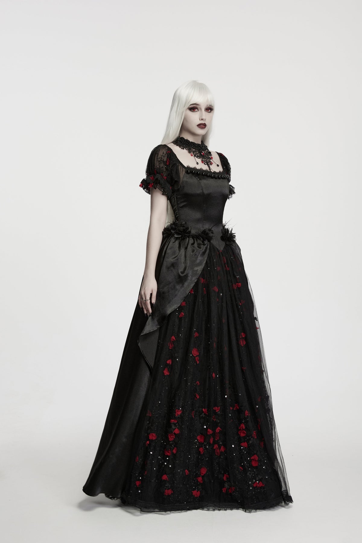 A model with platinum blonde hair wearing a long black gothic wedding gown with red rose embroidery, standing against a plain light grey background.