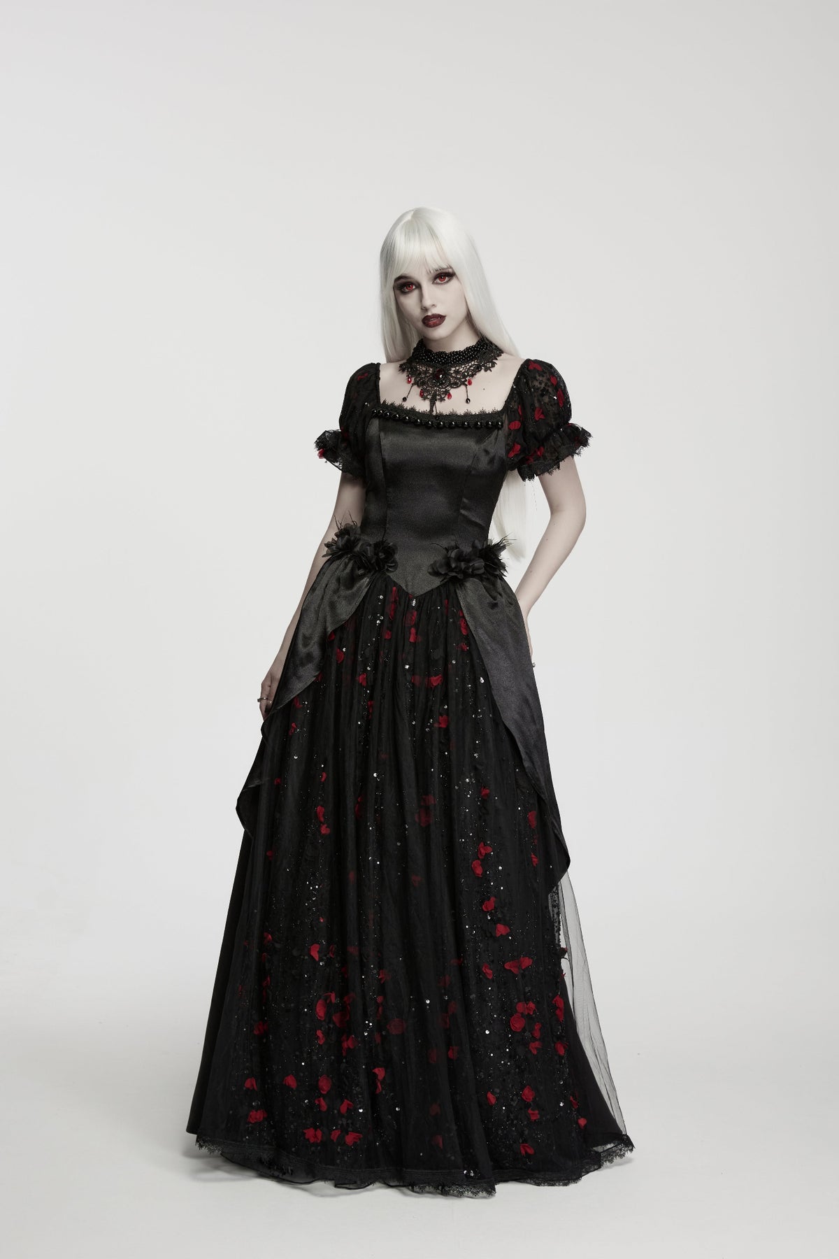 A gothic-style black ball gown with red rose embroidery, modeled by a woman with long white hair against a light grey studio background.