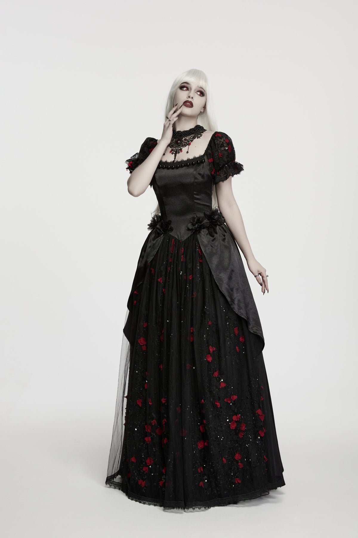 A gothic-style model with platinum blonde hair wearing a black long gown with red rose embroidery, a lace collar, and sheer layered sleeves, photographed against a seamless light gray studio background.