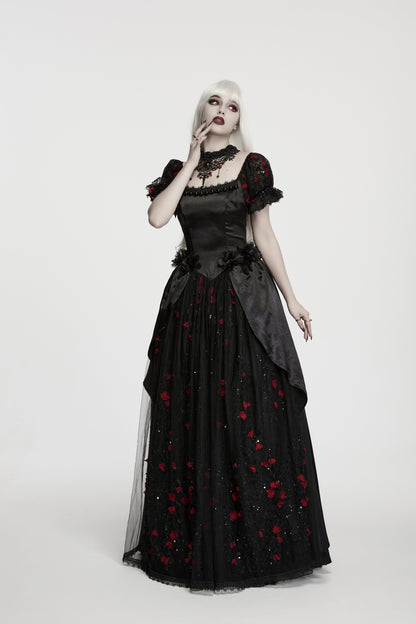 A gothic-style model with platinum blonde hair wearing a black long gown with red rose embroidery, a lace collar, and sheer layered sleeves, photographed against a seamless light gray studio background.