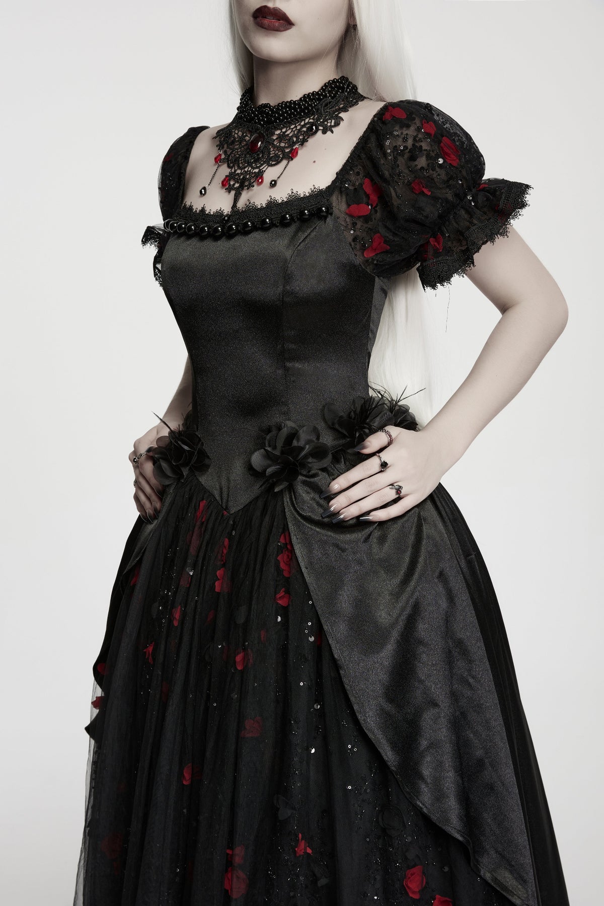 A woman modeling a black gothic-style dress with red rose embroidery, black lace puff sleeves, and a sheer tulle skirt, against a plain light grey background.