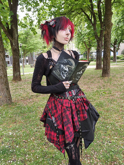 Valentinescorpse in a park wearing a gothic-inspired outfit with a red and black tartan skirt.