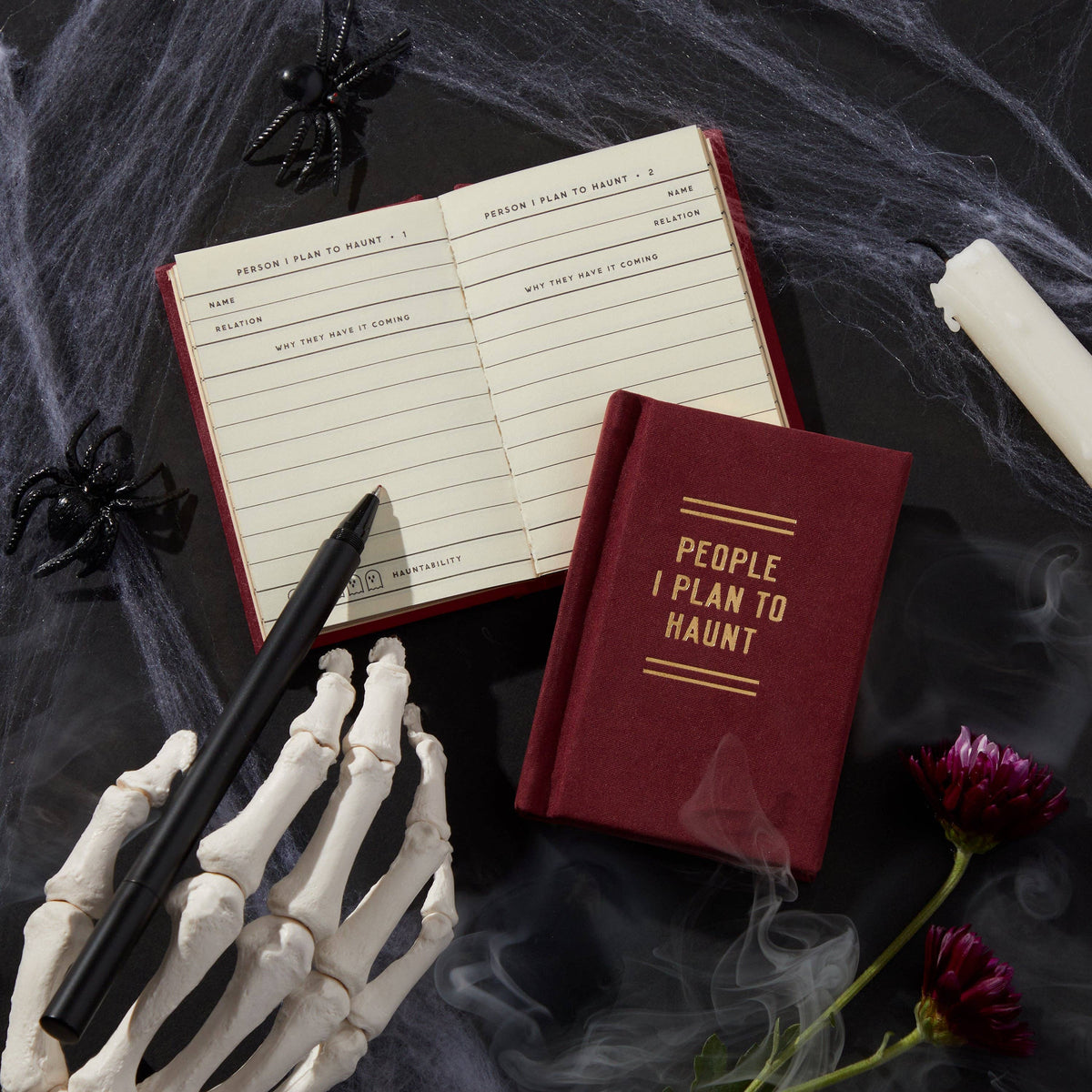The diary open and closed on dark fabric, beside a skeleton hand, a candle and dark flowers.