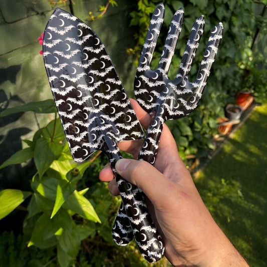 Hand holding a pair of black and white bat and moon patterned gardening tools with a garden background