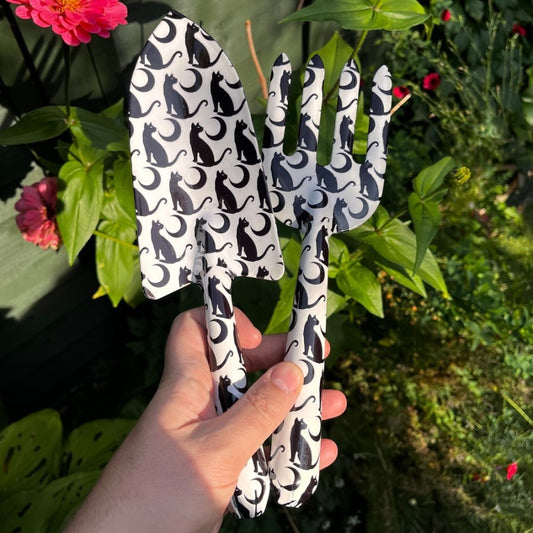 Gardening tools with black cat pattern held in front of green foliage and flowers